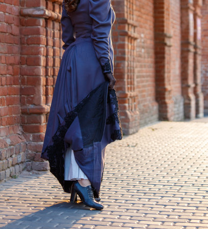 A woman in a long dress is standing on a brick sidewalk. The dress is blue and has a black lace trim. The woman is wearing black gloves and has her hands in her pocketsの写真素材