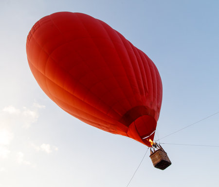 A red hot air balloon is flying high in the sky. The balloon is filled with hot air and is being carried by a string. The sky is clear and blue, and the sun is shining brightlyの写真素材