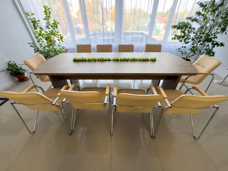 A long wooden table with a green plant in the middle and a bunch of chairs around it. The chairs are...