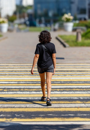A young woman walks across a crosswalk. She is wearing a black shirt and gray shorts. The crosswalk is yellow and whiteの写真素材