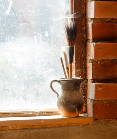 A vase with brushes in it sits on a window sill. The vase is made of clay and has a rustic appearance. The brushes are made of wood and are arranged in a neat rowの写真素材