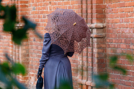A woman wearing a long dress and holding a brown umbrella. The umbrella is decorated with lace and...