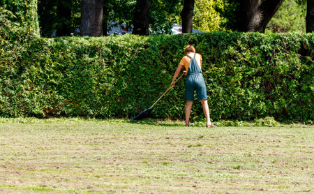 A woman in a green apron is walking through a hedge with a rake in her hand. The scene is peaceful and serene, with the woman taking her time to tend to the plantsの写真素材