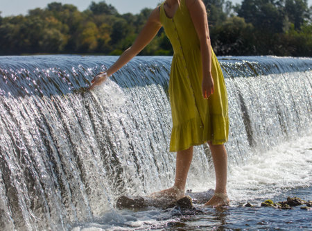 A woman in a yellow dress is standing on a rock in a stream. The water is flowing over the rock, creating a peaceful and serene atmosphere. The woman is enjoying the momentの写真素材