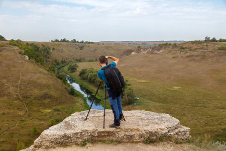A man with a camera is taking a picture of a river. The man is wearing a backpack and is standing on...