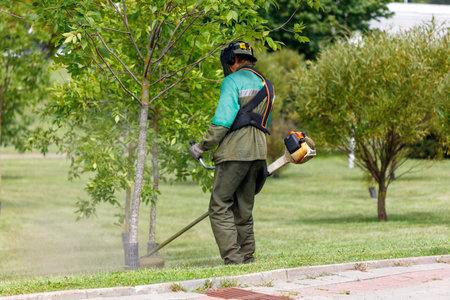 Worker using a grass trimmer to maintain a park, wearing protective gear and cutting grass near a...