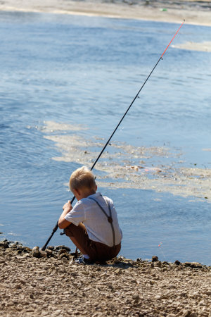 A young boy is fishing in a body of water. The boy is wearing a white shirt and brown shortsの写真素材