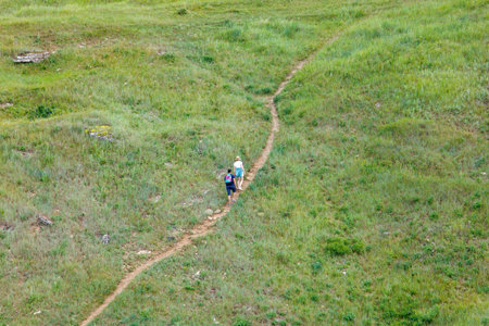 Two people walking on a dirt path in a grassy field. The path is narrow and winding, and the grass...