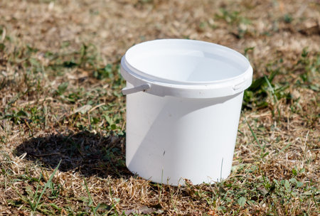 A white bucket is sitting on the ground in a field. The bucket is empty and the grass is dry