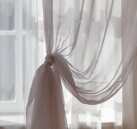 A white curtain is hanging in front of a window. The curtain is tied with a white ribbonの写真素材