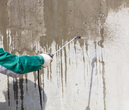 A man is spraying a wall with a spray gun. The wall is covered in paint and the man is wearing a green jacketの写真素材