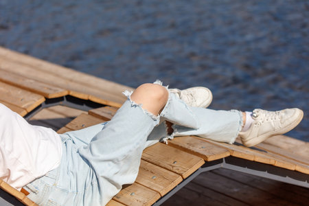 A person is laying on a wooden bench by a body of water. The person is wearing white shoes and blue...