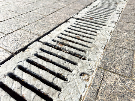A drain is shown in a gray color. The drain is located on a sidewalk. The drain is made of concreteの写真素材