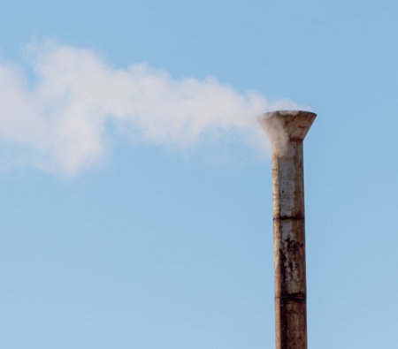 A tall, rusty pipe is spewing smoke into the sky. The sky is clear and blue, with no clouds in sight. The smoke is billowing out of the pipe, creating a sense of movement and energyの写真素材