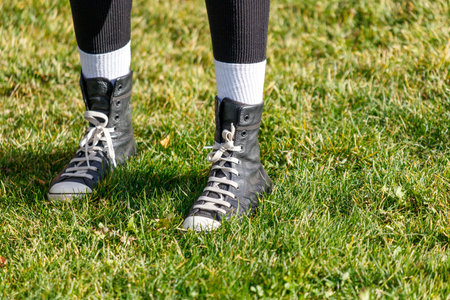 A person is standing in a field with their feet in the grass. They are wearing black shoes and white...