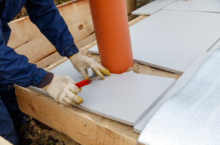A man is laying tiles on a floor. He is wearing a blue jacket and gloves. The tiles are gray and the floor is made of woodの写真素材