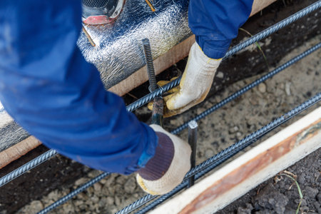 A man in a blue jacket is working on a construction site, holding a pair of pliers and bending a...