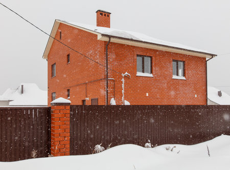 A brick house with a chimney and a fence in front of it. The house is covered in snowの写真素材