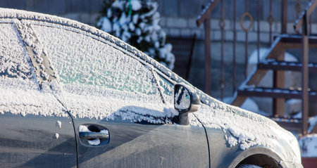 A car is covered in snow and has a frosted windshield. The car is parked in front of a house with a Christmas treeの写真素材