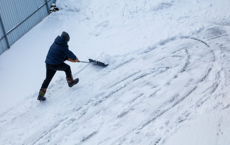 A man is shoveling snow off a driveway. The snow is piled up on the ground and the man is using a snow shovel to clear it. The scene is cold and snowy, and the man is working hard to clear the snowの写真素材