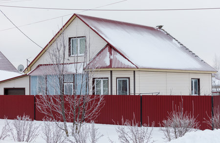 A house with a red fence and a red roof. The house is covered in snow. The house is surrounded by treesの写真素材