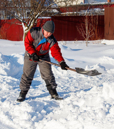 A man in a red jacket and gray pants is shoveling snow. He is wearing a hat and glovesの写真素材