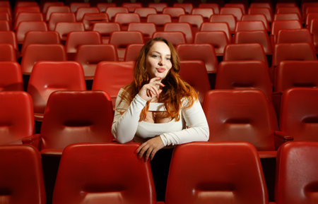 A woman sits in a red chair in a movie theater. She is wearing a white shirt and has her hands on her headの写真素材