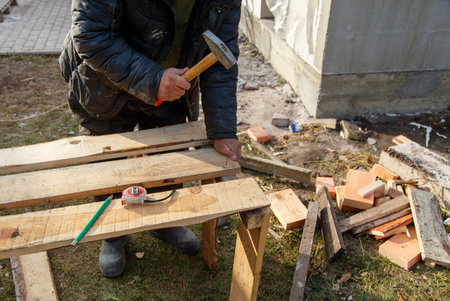 A man is hammering a piece of wood. The wood is on a table and there are other pieces of wood scattered around. The man is wearing a black jacket and he is focused on his taskの写真素材