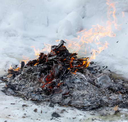 A pile of burnt wood with a lot of ash and snow on top. The fire is still burning and the ash is fallingの写真素材