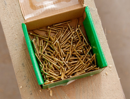 A box of screws sits on a table. The screws are gold and shiny. The box is green and has a lid