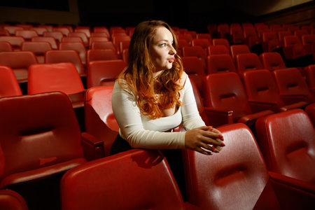A woman sits in a red chair in a movie theater. She is wearing a white shirt and has red hair