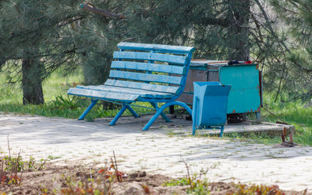 A blue bench sits in a park next to a trash can. The bench is empty and the trash can is full. The...