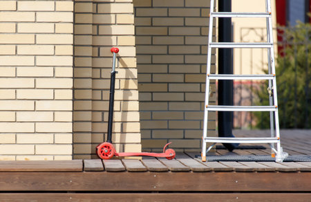 A red scooter is sitting on a wooden deck next to a ladder. The ladder is leaning against a brick wallの写真素材