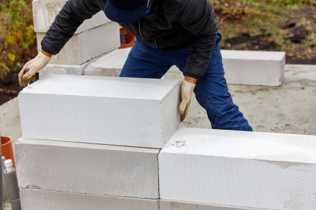 A man is laying bricks in a wall. He is wearing a black jacket and blue jeans. The bricks are white and he is using a trowel to lay themの写真素材