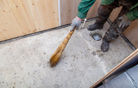 A man is sweeping a floor with a broom. The floor is dirty and the man is wearing a green shirt and black bootsの写真素材