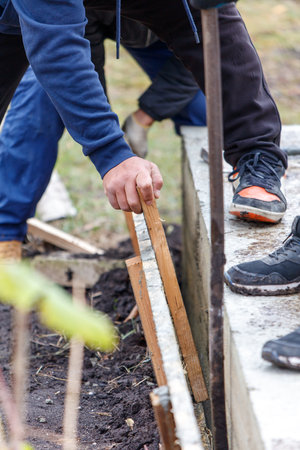 A man is working on a construction site, holding a piece of wood in his hand. The scene is set in a muddy area, with a few plants nearby. The man is focused on his taskの写真素材