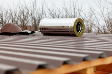A metal pipe is on a roof. The pipe is silver and has a green covering. The roof is brown