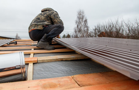 A man is working on a roof, fixing a piece of metal. Concept of hard work and dedication, as the man is crouching down to get the job done. Scene is focused and determinedの写真素材