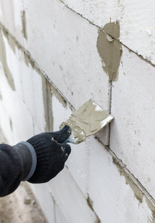 A person is painting a wall with a trowel. The wall is made of bricks and is white in color. The person is wearing a black glove and a black jacketの写真素材