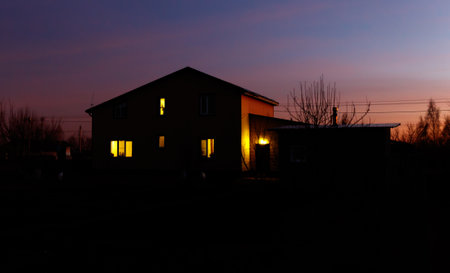 A house with a porch and a window is lit up at night. The house is surrounded by trees and the sky is a beautiful orange colorの写真素材