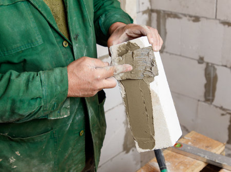 A man in a green jacket is holding a brick and applying cement to it. Concept of manual labor and construction workの写真素材