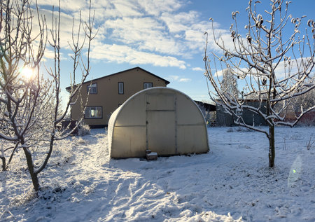 A greenhouse is covered in snow and is surrounded by trees. The house behind the greenhouse is also...