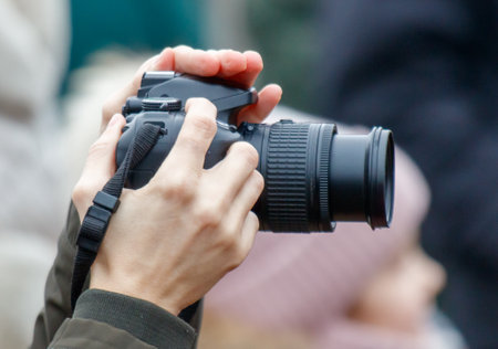 A person is holding a camera and taking a picture. The camera is black and has a strap. The person is wearing a green jacketの写真素材