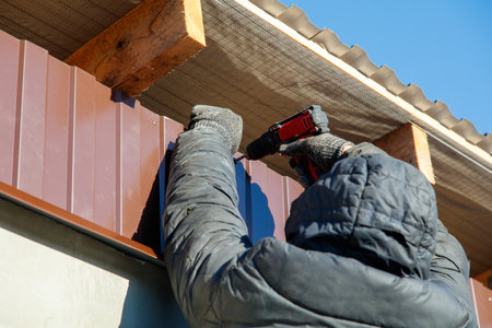 A man is working on a roof with a drill. He is wearing a black jacket and gloves
