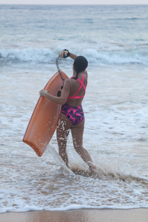 A woman is walking on the beach with a surfboard. She is wearing a pink and black bikiniの写真素材
