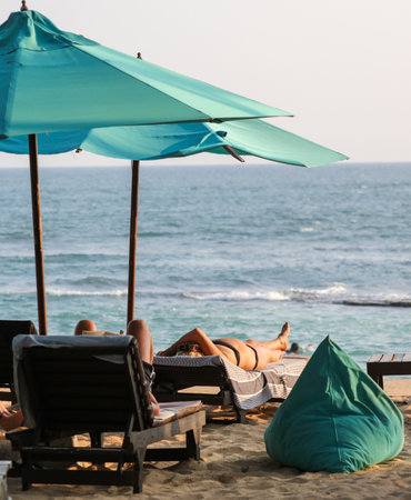 A woman is laying on a beach chair under a blue umbrella. The beach is empty and the ocean is calmの写真素材