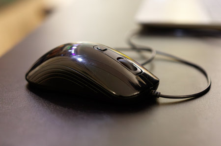 A black computer mouse is sitting on a desk. The mouse is connected to a cord. The mouse is not in...