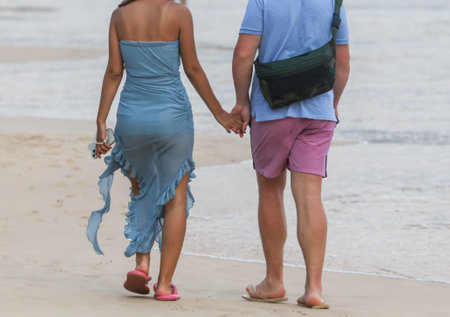 A man and woman are walking on the beach holding hands. The woman is wearing a blue dress and the man is wearing a blue shirt and pink shortsの写真素材