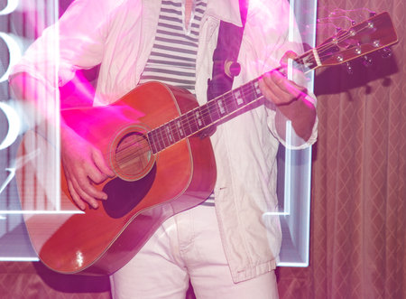 A man is playing a guitar in a room with a pink background. The guitar is a brown color and the man...