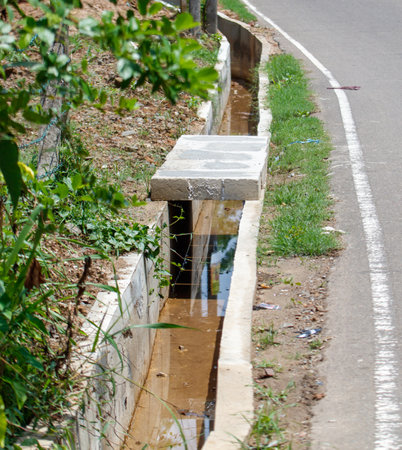 A dirty, muddy ditch runs along the side of a road. The water is murky and the grass is overgrownの写真素材